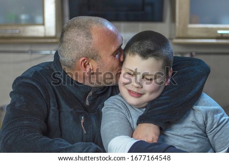 Dad and his son with autism sitting in kitchen at home. A man with short hair and bristle hugs his son and kisses him on the cheek. Royalty-Free Stock Photo #1677164548