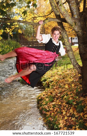 Playful barefoot young woman in a traditional dirndl swinging on the branches an autumn tree over a stream laughing with happiness /Playful young woman swinging on a tree