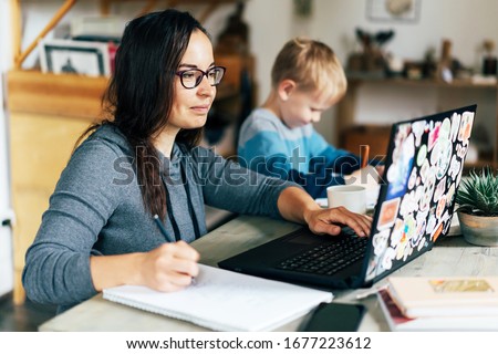 Concept of work from home and home family education. Mom and son are sitting at the desk. Business woman works on the Internet in a laptop, a child writes in a notebook. Royalty-Free Stock Photo #1677223612