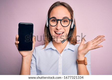 Beautiful call center agent woman working using headset holding smartphone holding screen very happy and excited, winner expression celebrating victory screaming with big smile and raised hands