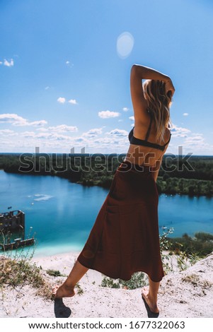 At a picturesque vantage point of Rummu quarry, a blond woman is standing in her bare feet, wearing a thigh high, auburn coloured dress, With her back turned away from the camera. Clear Water