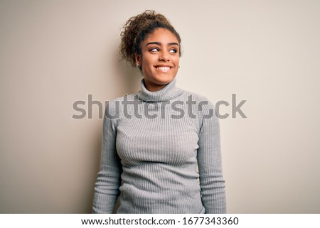 Beautiful african american girl wearing turtleneck sweater standing over white background looking away to side with smile on face, natural expression. Laughing confident.