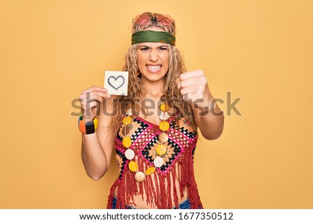 Beautiful blonde hippie woman wearing sunglasses and accessories holding reminder with heart annoyed and frustrated shouting with anger, yelling crazy with anger and hand raised
