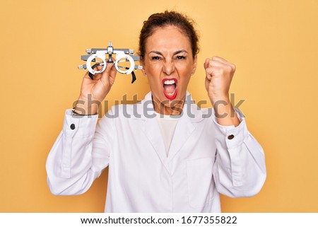 Middle age senior oculist woman holding optometrical glasses for vision correction annoyed and frustrated shouting with anger, crazy and yelling with raised hand, anger concept