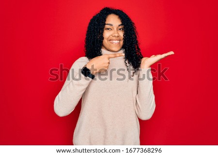 Young african american curly woman wearing casual turtleneck sweater over red background amazed and smiling to the camera while presenting with hand and pointing with finger.