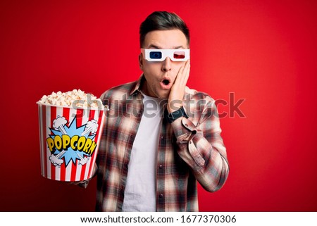 Young handsome caucasian man wearing 3d movie glasses and eating popcorn afraid and shocked, surprise and amazed expression with hands on face