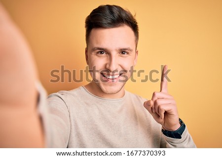 Young handsome caucasian man talking a selfie picture over yellow isolated background surprised with an idea or question pointing finger with happy face, number one