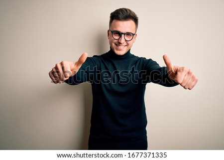 Young handsome caucasian man wearing glasses and casual sweater over isolated background approving doing positive gesture with hand, thumbs up smiling and happy for success. Winner gesture.