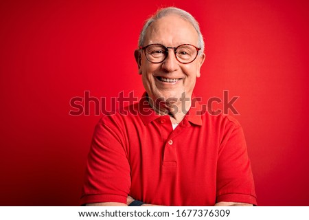 Grey haired senior man wearing glasses and casual t-shirt over red background happy face smiling with crossed arms looking at the camera. Positive person.