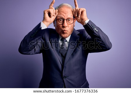 Grey haired senior business man wearing glasses and elegant suit and tie over purple background doing funny gesture with finger over head as bull horns