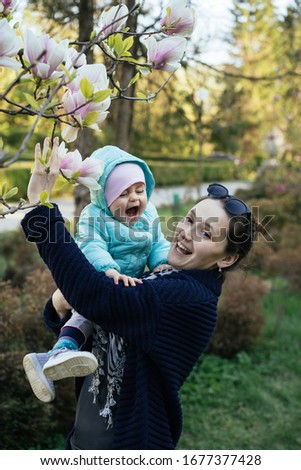 Spring family photo. Portrait of mother and baby in magnolia flowers. Blooming garden. Young stylish woman with toddler standing in park, enjoy the beauty and sniff pink flowers. Daughter cries