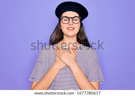 Young beautiful brunette woman wearing glasses and french beret over purple background smiling with hands on chest with closed eyes and grateful gesture on face. Health concept.