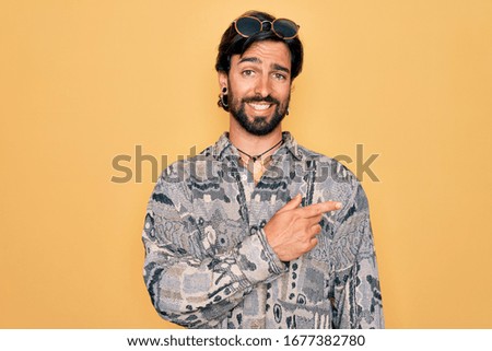 Young handsome hispanic bohemian man wearing hippie style and sunglasses cheerful with a smile of face pointing with hand and finger up to the side with happy and natural expression on face
