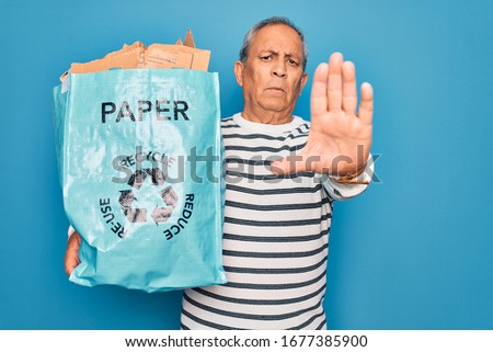 Senior grey-haired man recycling holding bag with cardboard to recycle over blue background with open hand doing stop sign with serious and confident expression, defense gesture