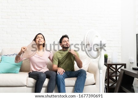 Latin heterosexual couple feeling hot while sitting in front of fan at home Royalty-Free Stock Photo #1677390691