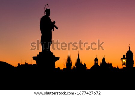 Silhouette of the statue of St. John of Nepomuk in Prague on the Charles Bridge at sunrise. You can also see the Týn Monastery and the Old Town Bridge Tower and the lighting of the Charles Bridge.