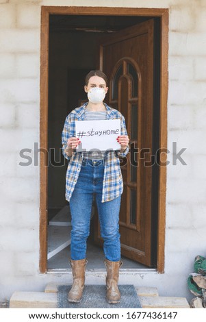 Coronavirus. Young woman in protection mask holding paper with hashtag Stay Home in her house with door background. Patient isolated to prevent infection of pandemic, epidemic Covid-19. Stop virus!