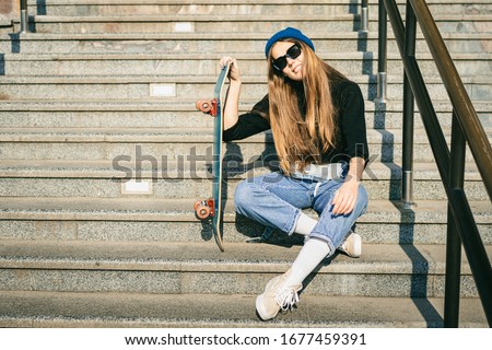 Stylishly dressed woman in blue denim jumpsuit posing with skateboard. Street photo. Portrait of girl holding skateboard. Lifestyle, youth concept. Leisure, hobby and skate in the city.