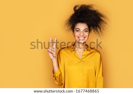 Smiling girl showing peace sign or V gesture with fingers. Photo of african american girl on yellow background Royalty-Free Stock Photo #1677468865