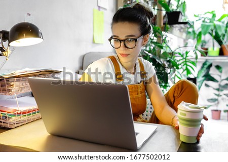 Young female gardener in glasses using laptop, communicates on internet with customer in home garden/greenhouse, hold reusable coffee/tea mug.Cozy office workplace, remote work, stay home concept Royalty-Free Stock Photo #1677502012