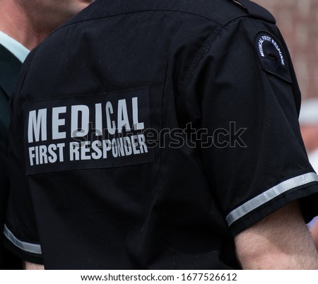 An adult male stands next to a man wearing an emergency medical first responder uniform. The short sleeve shirt is a uniform of navy blue with grey reflective letters. There are reflective arm bands. Royalty-Free Stock Photo #1677526612