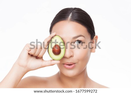 Young latino woman holding avocado with playful face on a white background