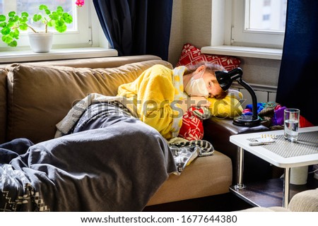 Photo of an adult woman with a viral infection, lying at home on a sofa in a medical mask. Sad mood, sadness, longing