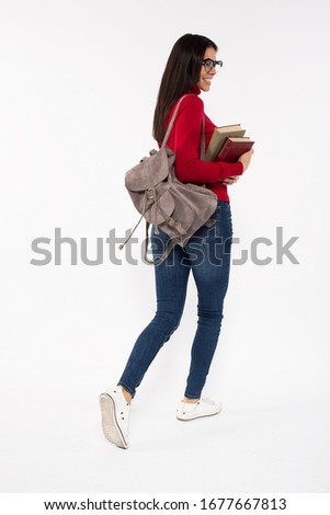 Photo of young female student walking with a backpack, looking over her shoulder and smiling, holding books in her hands, wearing casual outfit and reading glasses, isolated on white background