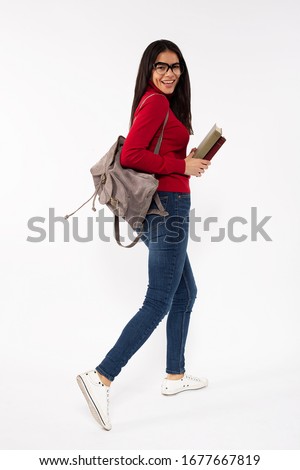 Photo of young female student walking with a backpack, looking over her shoulder and smiling, holding books in her hands, wearing casual outfit and reading glasses, isolated on white background