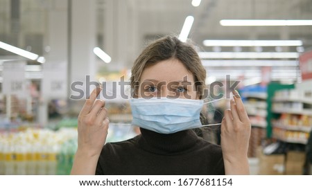 A young woman puts on a medical mask in a grocery supermarket close-up and looks at the camera, protection from coronavirus, shopping in a store to wait out the quarantine. Royalty-Free Stock Photo #1677681154