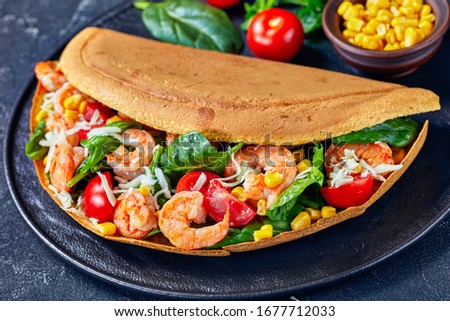 Singapore thick pancake with shrimps, spinach leaves, corn, cherry tomatoes, shredded white cheese on a black plate with ketchup and parsley on a dark concrete background, close-up, horizontal