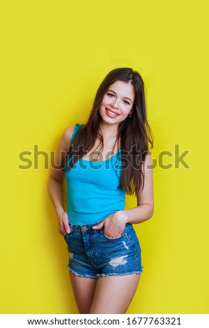 Cute young woman in blue T-shirt and denim shorts posing over yellow background.
