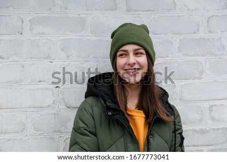 Portrait of a beautiful smiling girl with braces in a yellow sweater and khaki hat, which stands near a white brick wall. The concept of emotions and copy space Royalty-Free Stock Photo #1677773041