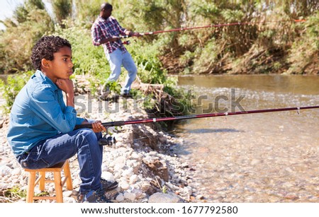 Positive African man and little boy standing near river and fishing with rods
