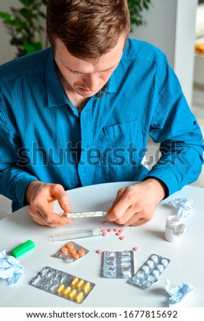 Shot of a young man holding a thermometer and many pills on a table at home. Actual problem, cold and chill, background for medical design