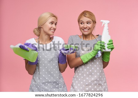Studio photo of pretty young white-headed females keeping bottles with household chemicals and looking excitedly at camera. Housework and housekeeping concept