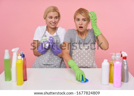 Confused young white-headed cleaning ladies wearing rubber gloves while making spring cleaning with household chemicals, isolated over pink background