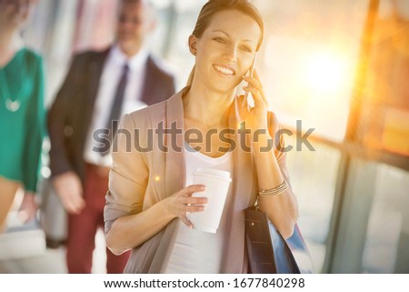 Portrait of young attractive businesswoman talking on smartphone and holding cup of coffee while walking against colleague