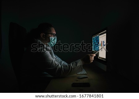 Stock photo of doctor looking at medical results on computer and pointing at them. He wears a white coat and a protective mask.