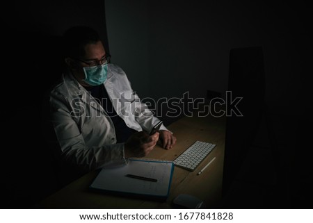 Stock photo of doctor looking at his smartphone in the office. He wears a white coat and a protective mask.