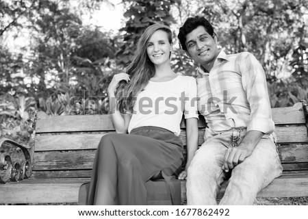 Happy multi ethnic couple smiling and sitting on wooden bench in love at peaceful green park