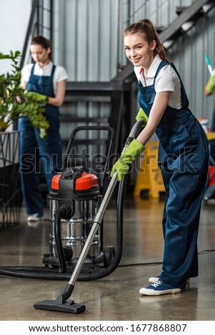 selective focus of smiling cleaner vacuuming floor near colleague cleaning plants