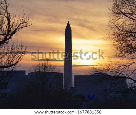 Photo shows Washington monument on a beautiful sunset evening where the sky is in golden color and the monument is dark. There are a few public museum around the monument.