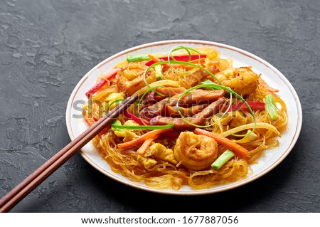 Singapore Mei Fun in white plate on dark slate background. Singapore Noodles is chinese cuisine dish with rice noodles, prawns, char siu pork, carrot, red onion, napa cabbage. Chinese food. Royalty-Free Stock Photo #1677887056