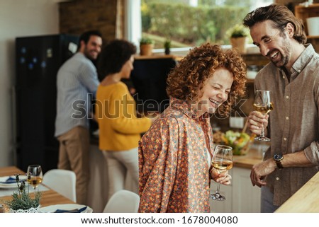 Young couple having fun and laughing while drinking wine in the kitchen. Their friends are preparing food in the background. 