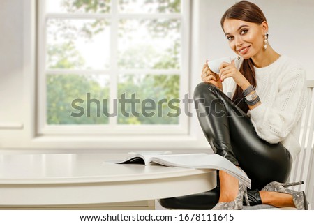 Slim young woman on chair with empty white wooden table of free space for your decoration.Coffee in white cup and blurred window sill in home interior.Spring time and sunny warm day.
