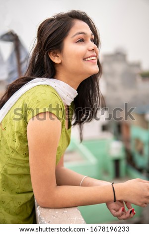 Beautiful happy Indian late teen girl enjoying fresh air in outdoor at day time. She is looking up and thinking with smile on her face. She is wearing traditional dress salwar Kameez and Dupatta.  Royalty-Free Stock Photo #1678196233