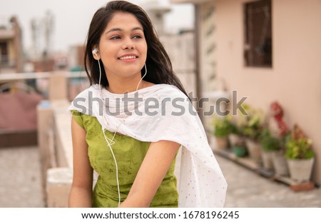 Beautiful happy Indian late teen girl listening music on smartphone through earphones and enjoying fresh air in outdoor on roof at day time. She is wearing traditional dress salwar Kameez and Dupatta. Royalty-Free Stock Photo #1678196245
