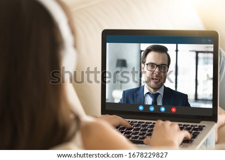 Pc screen view over woman shoulder, female wear headphones sitting on sofa make video call have distant communication using videoconference app working from home to prevent corona virus spread concept Royalty-Free Stock Photo #1678229827