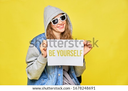 Stylish Caucasian girl in jeans and glasses 8 bit on a yellow background. Holding a piece of white paper in her hands with the inscription be yourself. Close up.